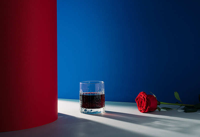 由一杯飲料和一朵紅玫瑰組成的創意浪漫安排。藍色和紅色的背景與光和陰影。最小的愛的概念。情人節的靈感。圖片下載