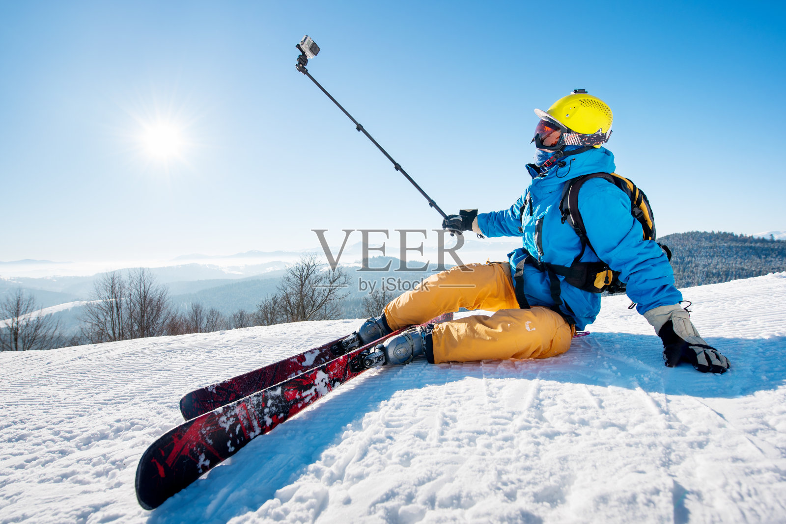 滑雪者坐在滑雪坡上自拍，使用自拍桿休息放松的極端娛樂生活方式活動技術概念。藍色的天空與太陽和冬天的森林背景照片攝影圖片