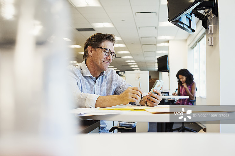 一个男人坐在办公室的桌子旁，看着手机。图片素材