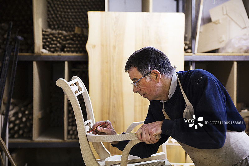 一个男人站在一个木工车间，在一把木椅上工作，用铅笔在扶手连接处做标记。图片素材