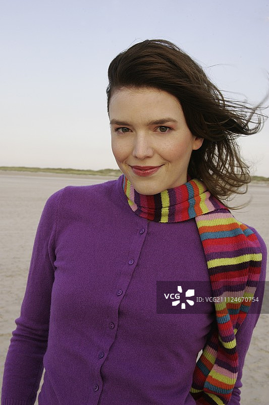 漂亮的金发女人穿着紫色的毛衣和条纹围巾站在海滩上，微笑图片素材