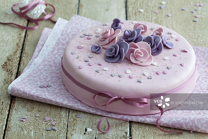 一个优雅的粉红色生日蛋糕，上面有软糖花和色带，放在一张质朴的木桌上图片素材