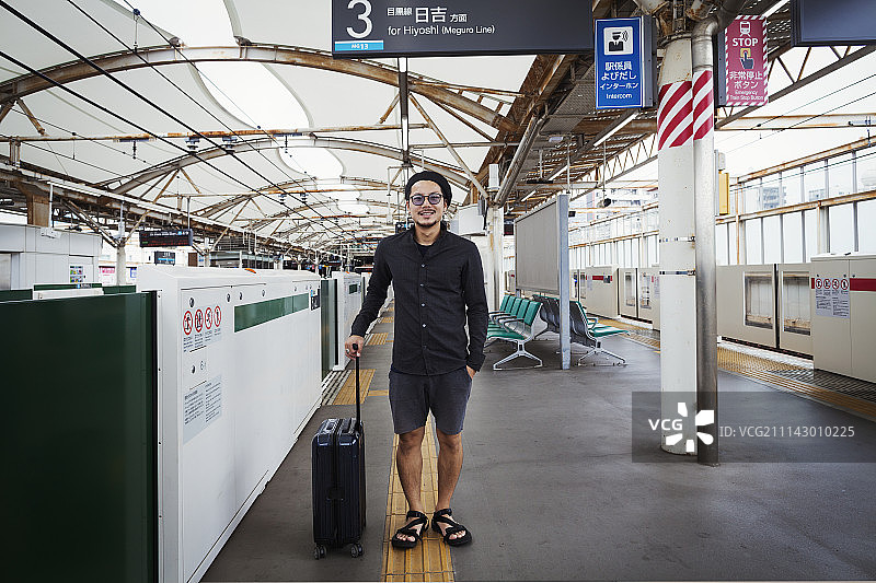 一名微笑的男子带着带轮子的手提箱站在地铁站的站台上，东京通勤者。图片素材