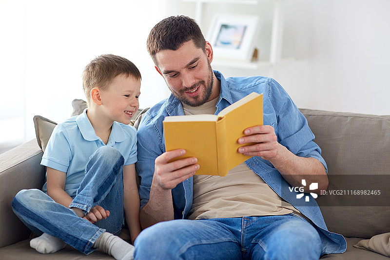 家庭、童年、父亲、休闲、人的概念——快乐微笑的父亲和儿子在家里沙发上看书的肖像。快乐的父子俩坐在家里的沙发上看书图片素材