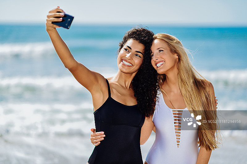 两个女人在海滩上用智能手机自拍。两名年轻女子在热带海滩上穿着泳衣用智能手机自拍。图片素材