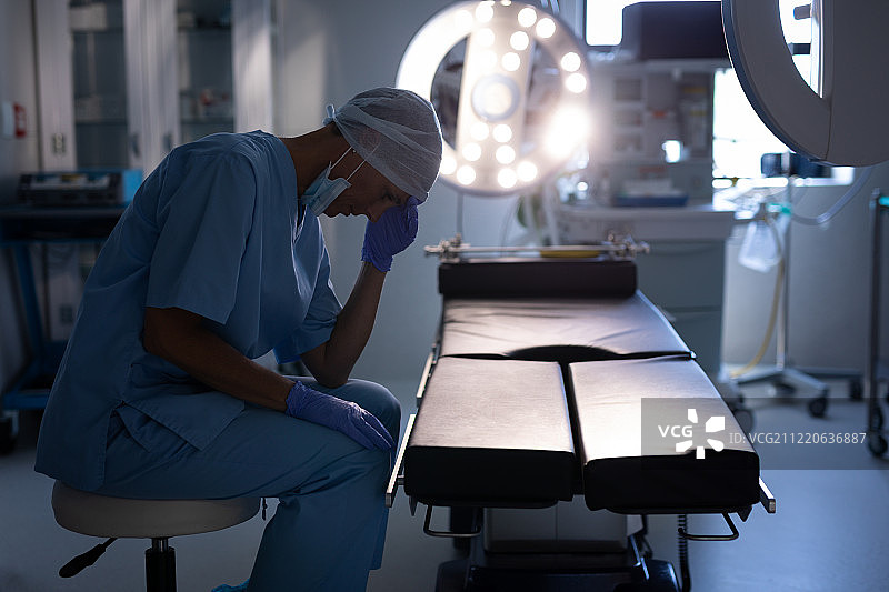 一位紧张的白种女外科医生手捧着头坐在医院的手术室里的侧视图图片素材