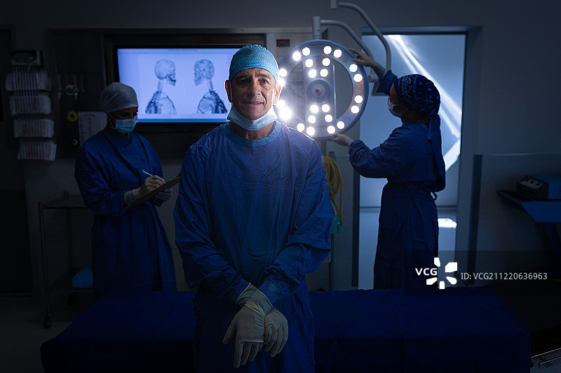 一名白人男性外科医生在手术室里看着摄像机，同事们在后面工作图片素材