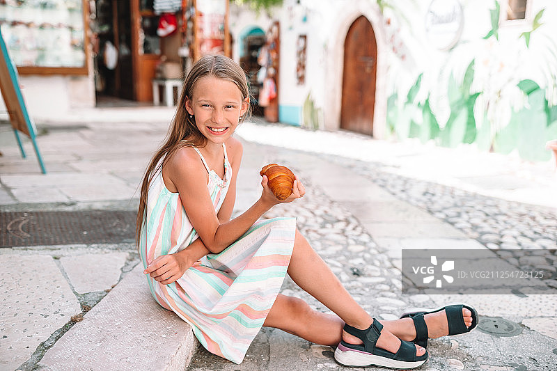 小女孩在欧洲村庄的老街上吃羊角面包图片素材