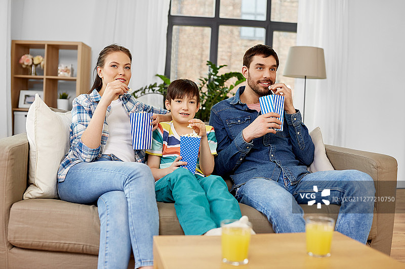 家庭，休闲和人的概念——快乐的妈妈，爸爸和儿子在家里吃爆米花和看电视。幸福的家庭和爆米花在家里看电视图片素材
