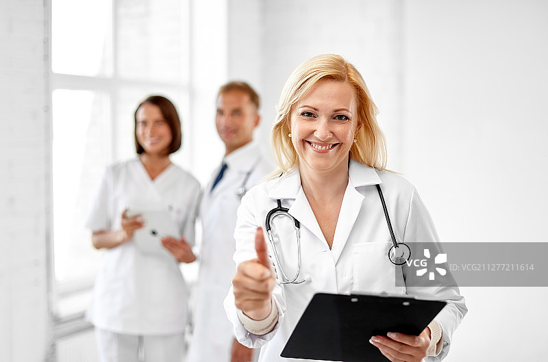 医疗保健，医学和职业概念-微笑女性中年医生与剪贴板和听诊器显示大拇指在医院。微笑的医生在医院竖起大拇指图片素材