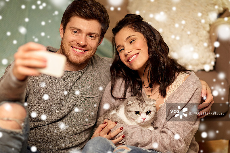 圣诞节，科技和人的概念-幸福的夫妇和猫在家里用智能手机自拍在雪中。幸福的夫妇在家里用智能手机自拍图片素材