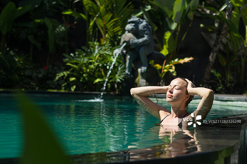性感的年轻女人放松在室外温泉无限游泳池与郁郁葱葱的热带图片素材