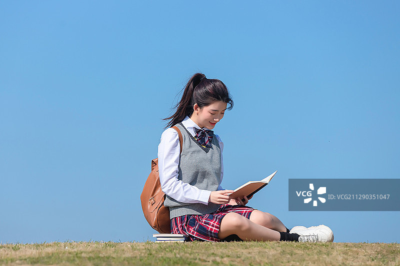 蓝色天空背景下，坐在草地上的亚洲女学生图片素材