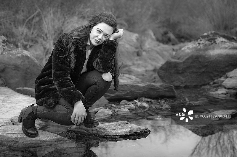 微笑的年轻女子蹲在森林河边的岩石上的肖像，蒙特莫索，西班牙图片素材