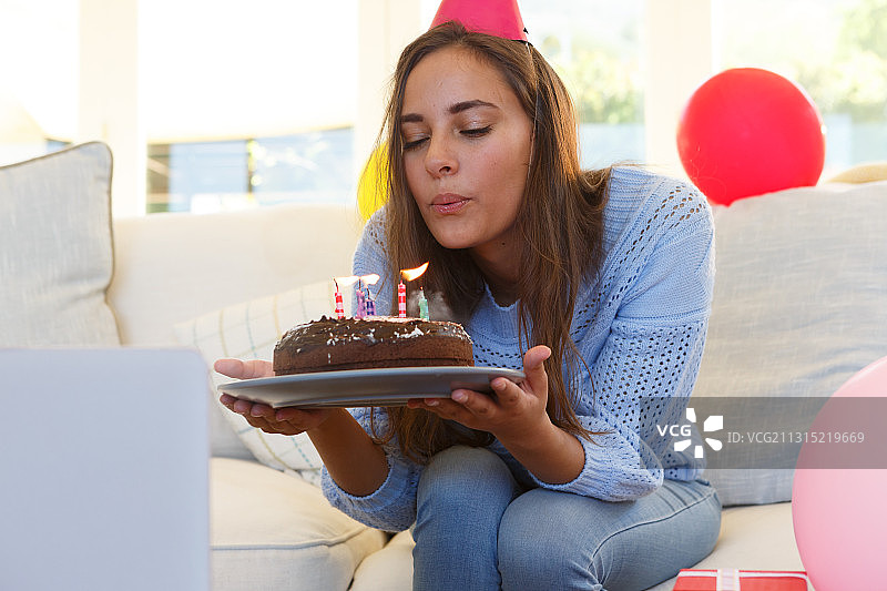 一个白种女人戴着派对帽在做生日视频电话吹灭蛋糕上的蜡烛图片素材