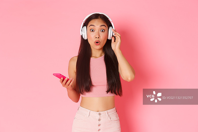 一名年轻女子在粉色背景下使用手机时戴着耳机的肖像图片素材