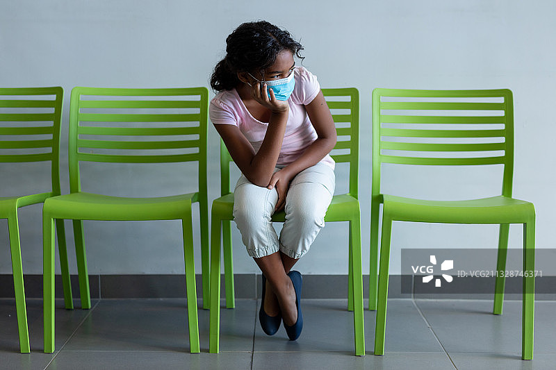 混血女孩戴着口罩坐在医院候诊室的绿色椅子上图片素材