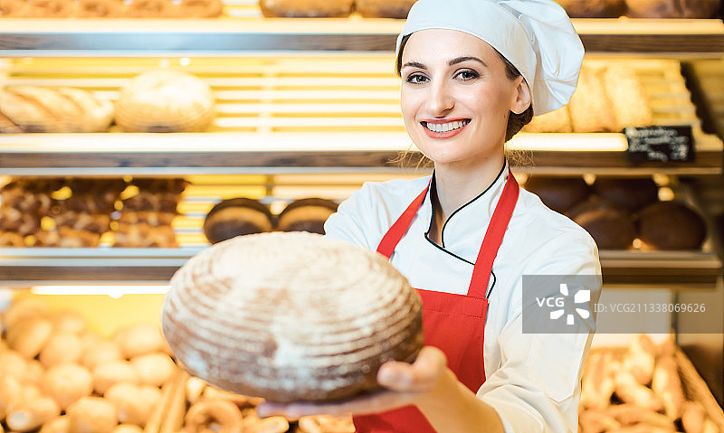 在面包店里，戴着围裙的女售货员正对着镜头展示新鲜的面包图片素材