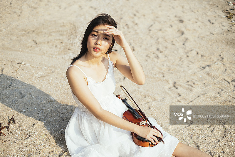 黑色长发的小提琴手坐在沙滩上，小提琴放在她的屁股上，她举起手来保护她的eY不被阳光照射。图片素材