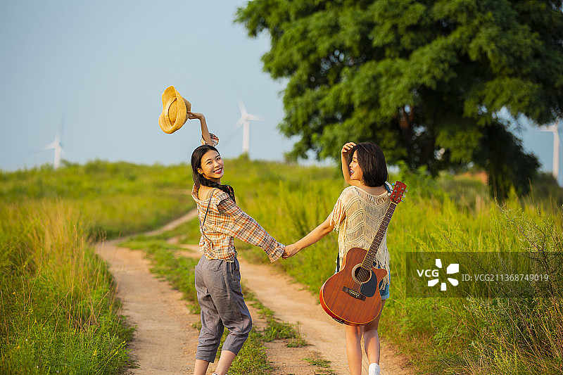 年轻闺蜜户外郊游，背着吉他手牵手走在乡村的小路上图片素材