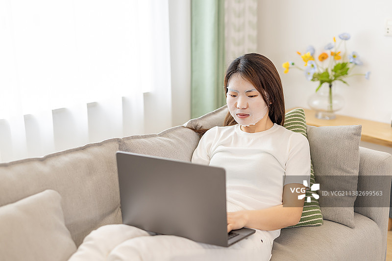 一位亚洲年轻女性在沙发上躺着敷面膜用笔记本电脑办公图片素材