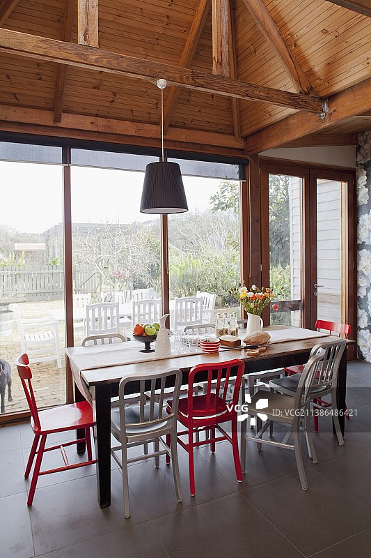 红色和白色的椅子围绕着玻璃墙旁边的质朴的木制桌子，下面是裸露的木制屋顶结构图片素材