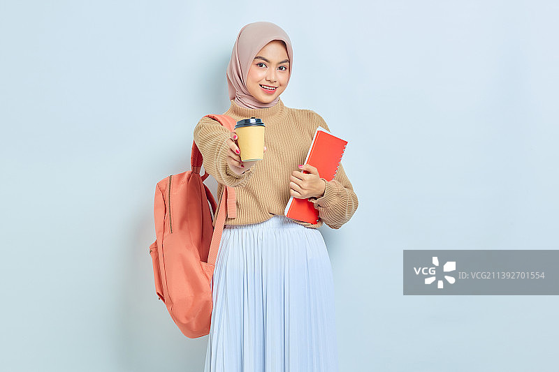 微笑的年轻亚洲穆斯林女学生在棕色毛衣，拿着书和杯咖啡图片素材