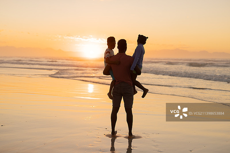 非洲裔美国年轻人的后视图拾起儿子和女儿在海滩上的天空在日落图片素材