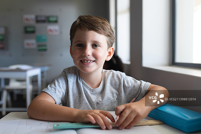 微笑的白人小学男孩的肖像，书和铅笔坐在课桌在教室里图片素材