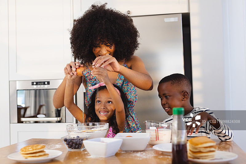 留着爆炸头的多种族母亲一边和女儿在碗里打鸡蛋，一边在桌上做早餐图片素材