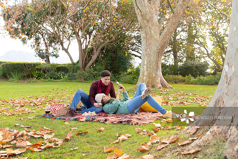 幸福的混血夫妇在地毯上野餐，在秋天的花园里玩智能手机图片素材