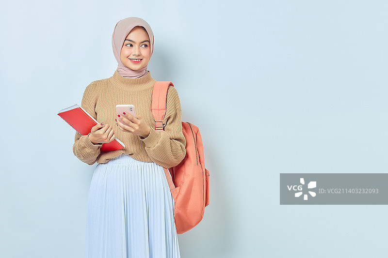 微笑的年轻亚裔穆斯林女学生，穿着棕色毛衣，拿着书和手机图片素材