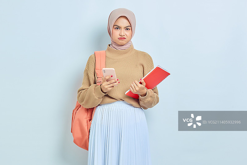 震惊的年轻亚裔穆斯林女学生，穿着棕色毛衣，拿着书和手机图片素材