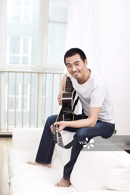 一个坐在白色沙发上弹吉他的青年男士图片素材