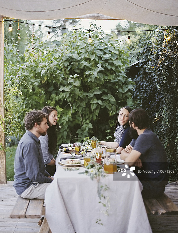 四个人坐在花园里的一张桌子旁。在白色桌布上摆放餐具和装饰品。两男两女。图片素材
