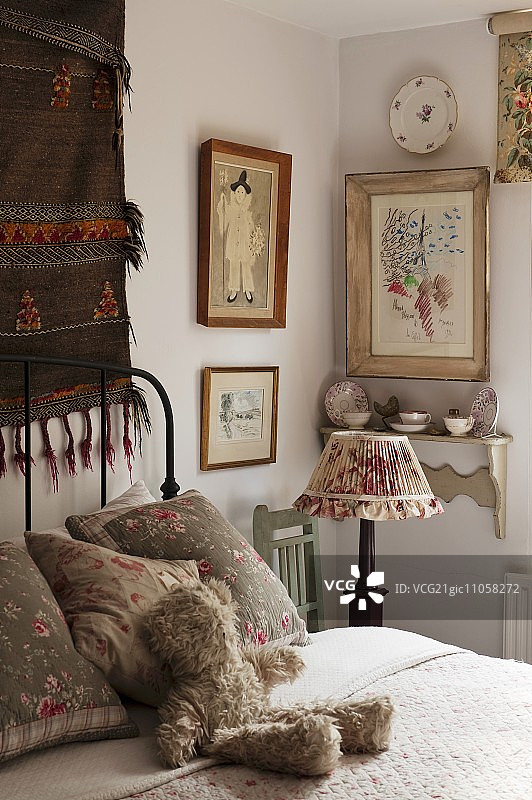 女性卧室的一角，木椅上怀旧的床头灯后面的壁架上挂着相框和有图案的瓷器;可爱的泰迪熊在舒适的床上等待图片素材