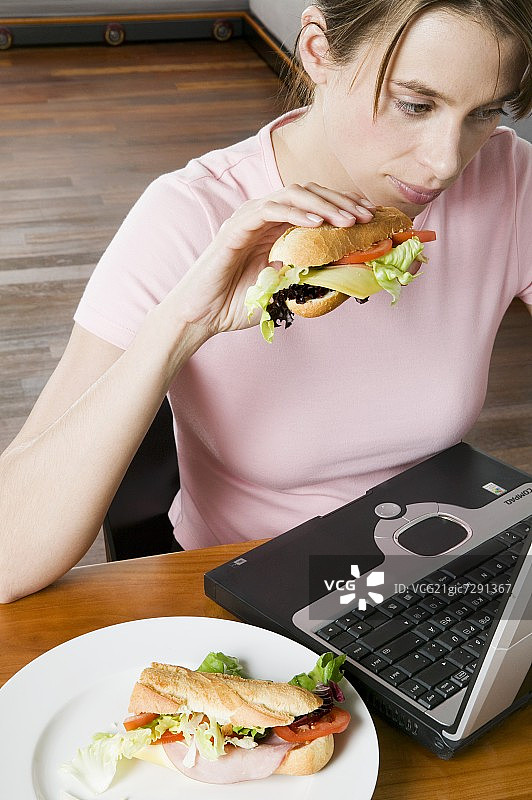 年轻女子一边吃三明治一边在电脑前工作图片素材