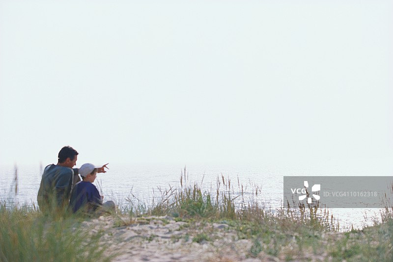 父子俩坐在面朝大海的沙丘上图片素材