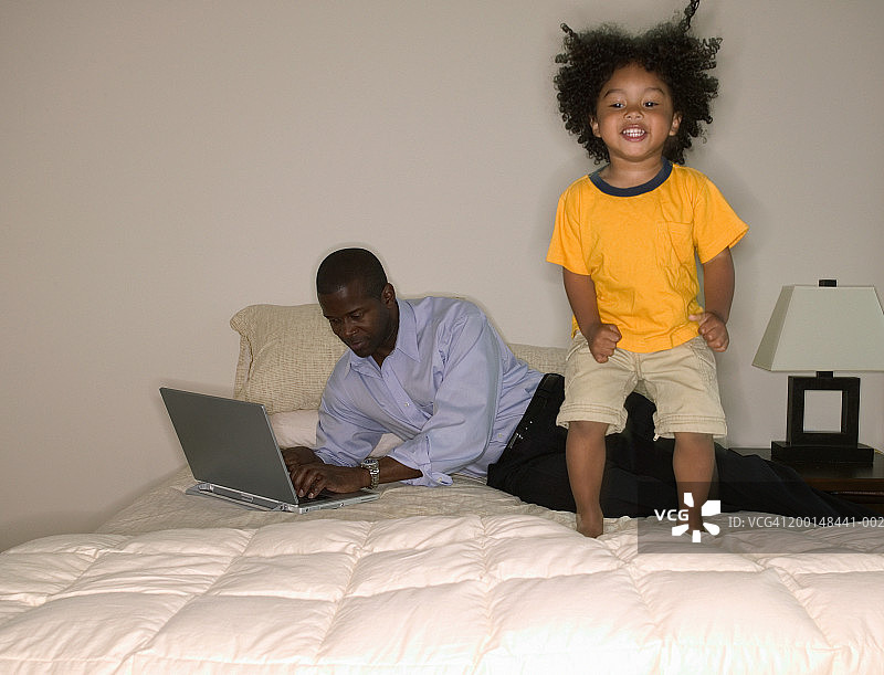 父亲在床上使用笔记本电脑，儿子(21-24个月)在床上跳图片素材