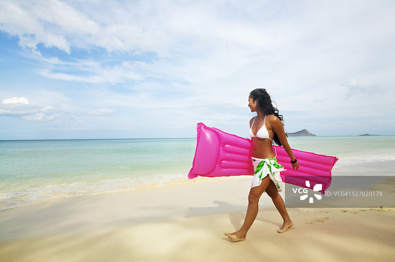 年轻女子背着充气床垫在海滩上散步图片素材