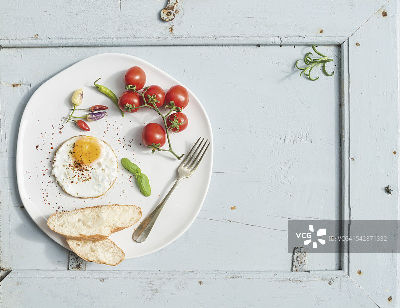 早餐。煎蛋，面包片，樱桃番茄，辣椒和香草放在白色陶瓷盘子上，浅蓝色的木制背景，俯视图。图片素材