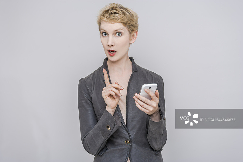 肖像的金发女子摇手指和智能手机在前面的灰色背景图片素材