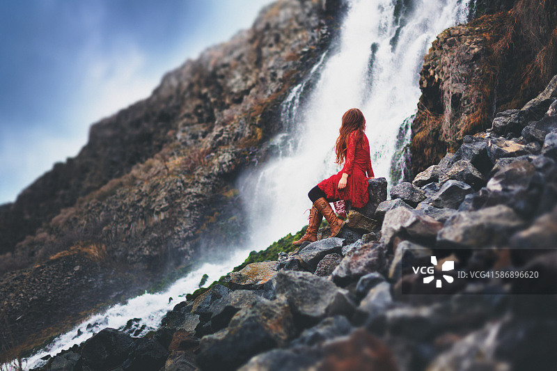 后视图的女人在红色蕾丝裙子和靴子坐在瀑布流过熔岩悬崖图片素材