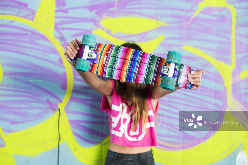 十几岁的女孩站在墙上涂鸦隐藏在五颜六色的滑板后面图片素材