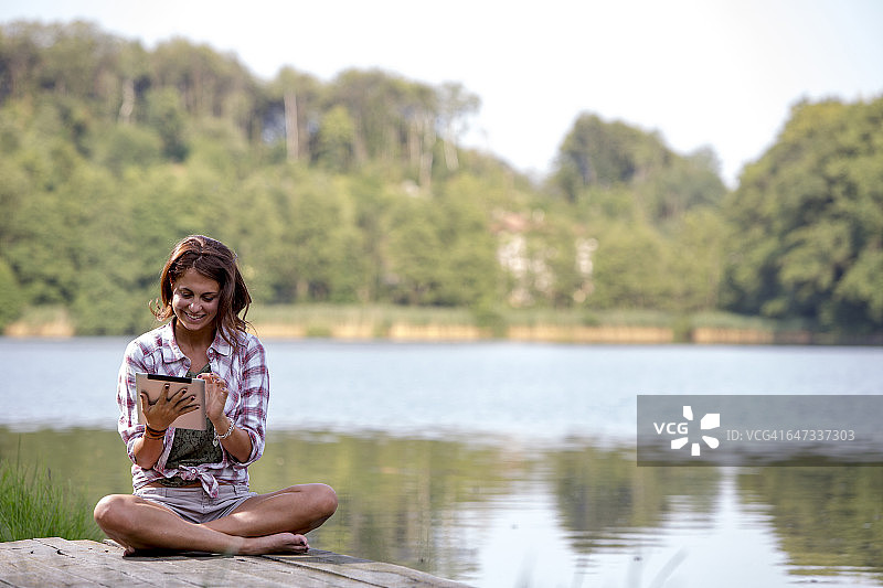 一个年轻女子拿着平板电脑坐在湖边的防波堤上图片素材