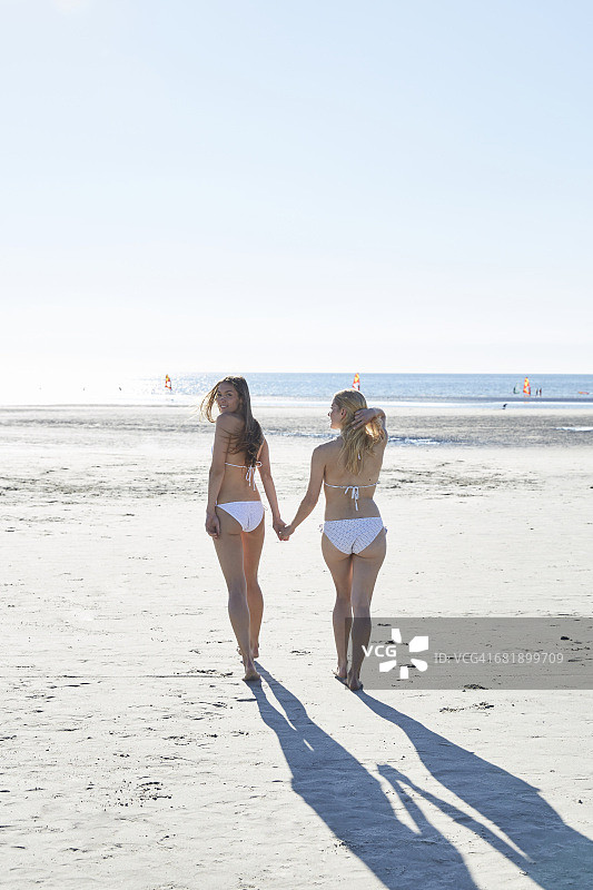 两个穿着比基尼的女性朋友在海滩上手拉手散步图片素材
