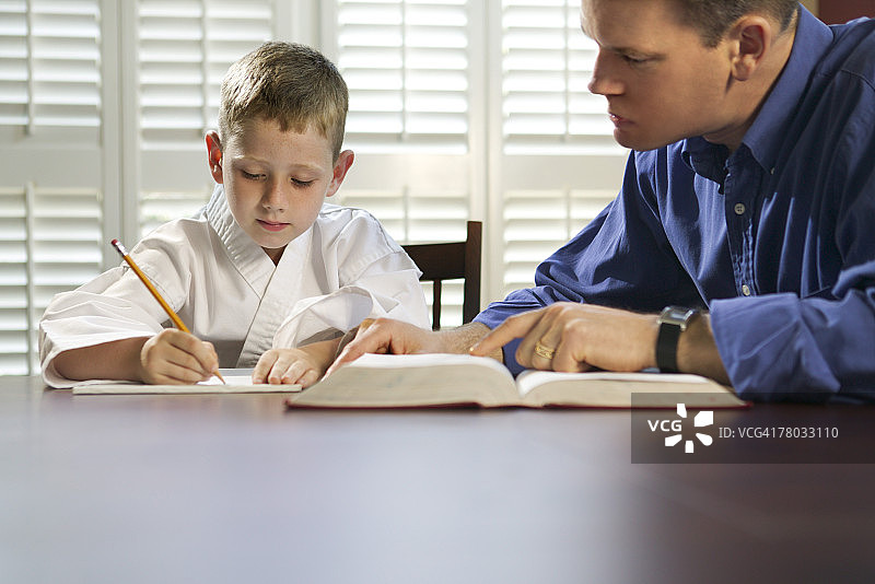 一个穿着武术服的男孩在写作业，他的爸爸在一旁看着他图片素材