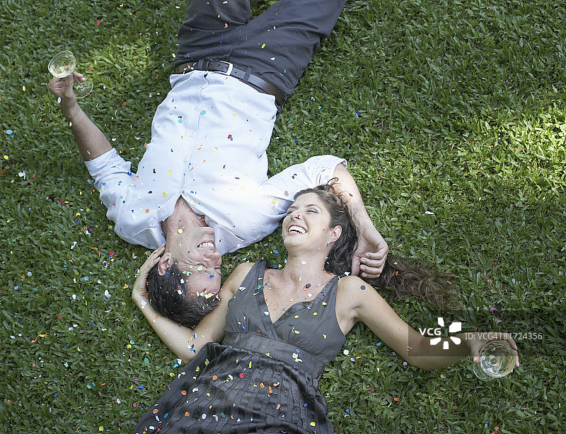 一对夫妇躺在户外草地上，喝着白葡萄酒，撒着五彩纸屑开怀大笑图片素材