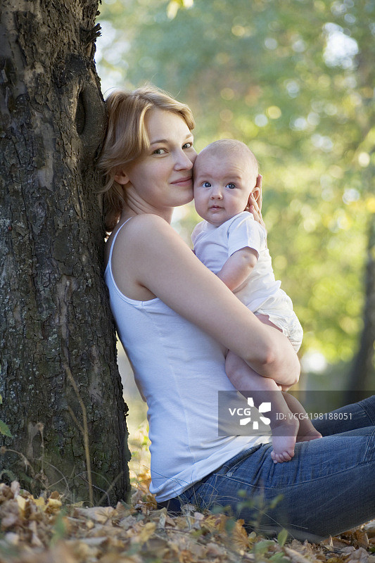 一位母亲坐在一棵树旁亲吻她的孩子，看着镜头图片素材