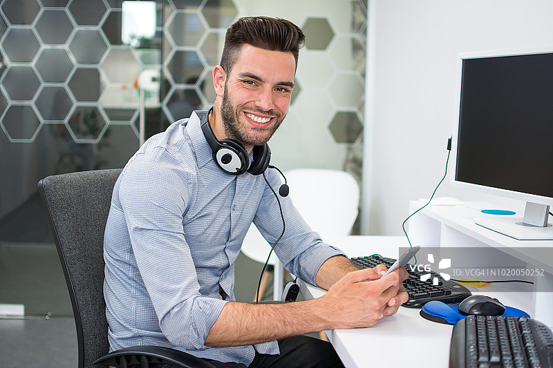 微笑英俊的接线员与耳机坐在附近的电脑在他的工作和使用电话。图片素材